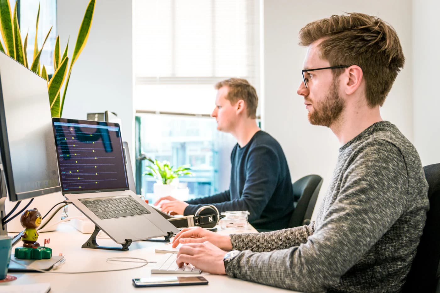Twee mensen aan een bureau vol beeldschermen, één laptop toont regels programmeercode